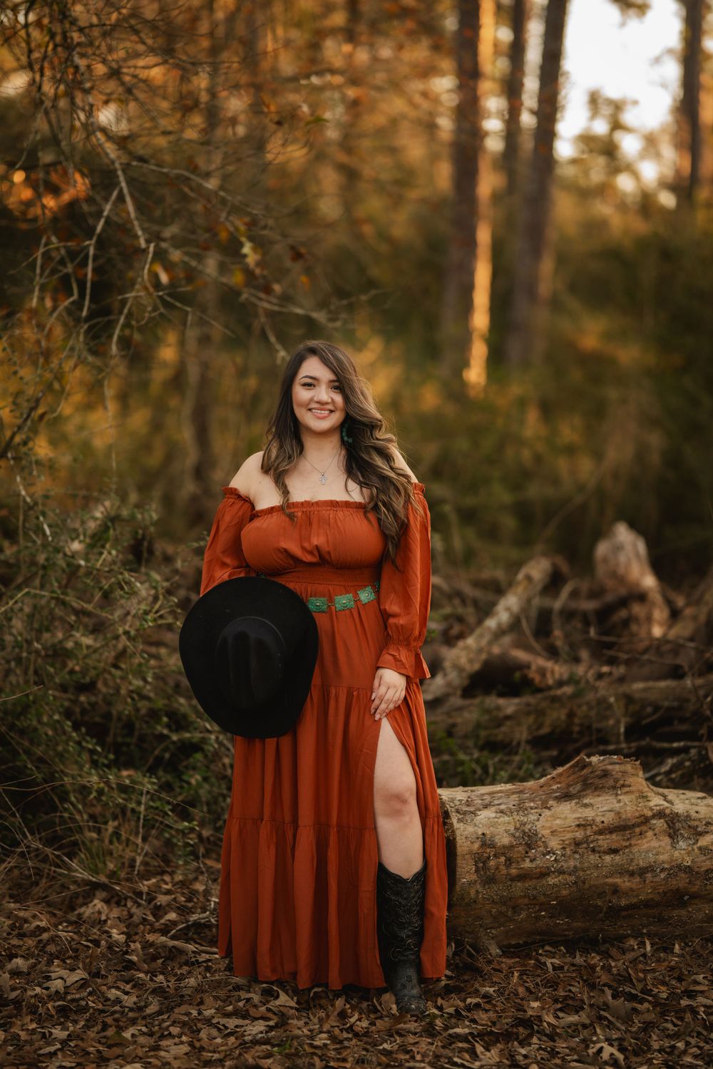 A rustic autumn forest portrait features a long flowing rust-colored dress and black hat against golden sunlit trees.