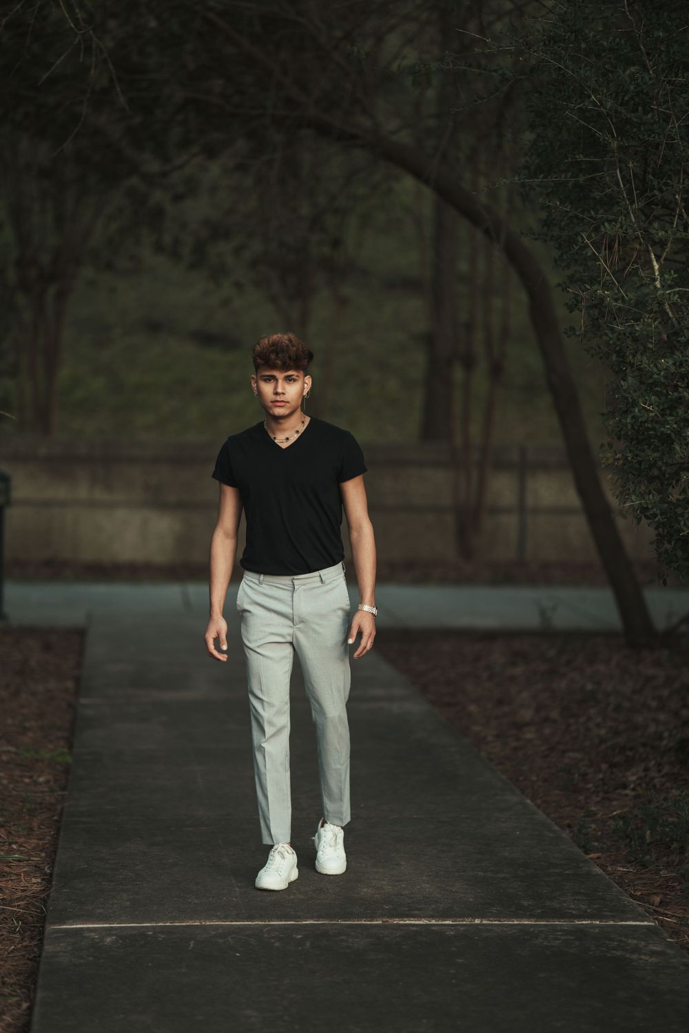 A figure in a black t-shirt and khaki pants walks along a tree-lined walkway at dusk.