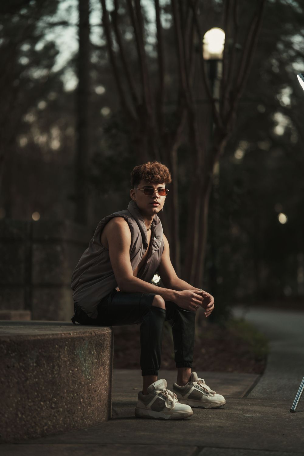 A person sits pensively on a concrete ledge at dusk wearing a tank top and sneakers in a park setting.