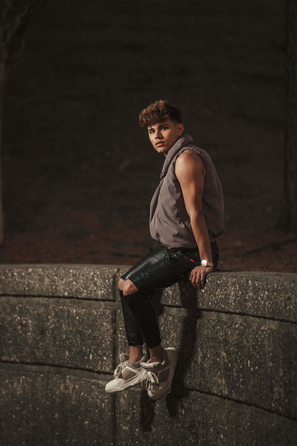Person in a gray sleeveless top and dark pants sits on concrete wall against dark background in moody lighting.