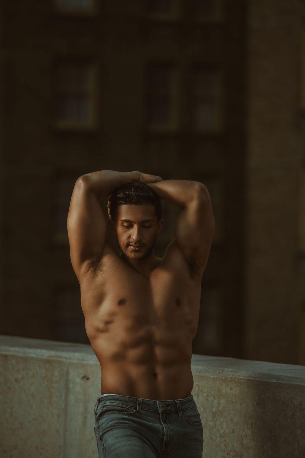 Artistic low light portrait of shirtless muscular person posing against dark urban backdrop with dramatic shadows.