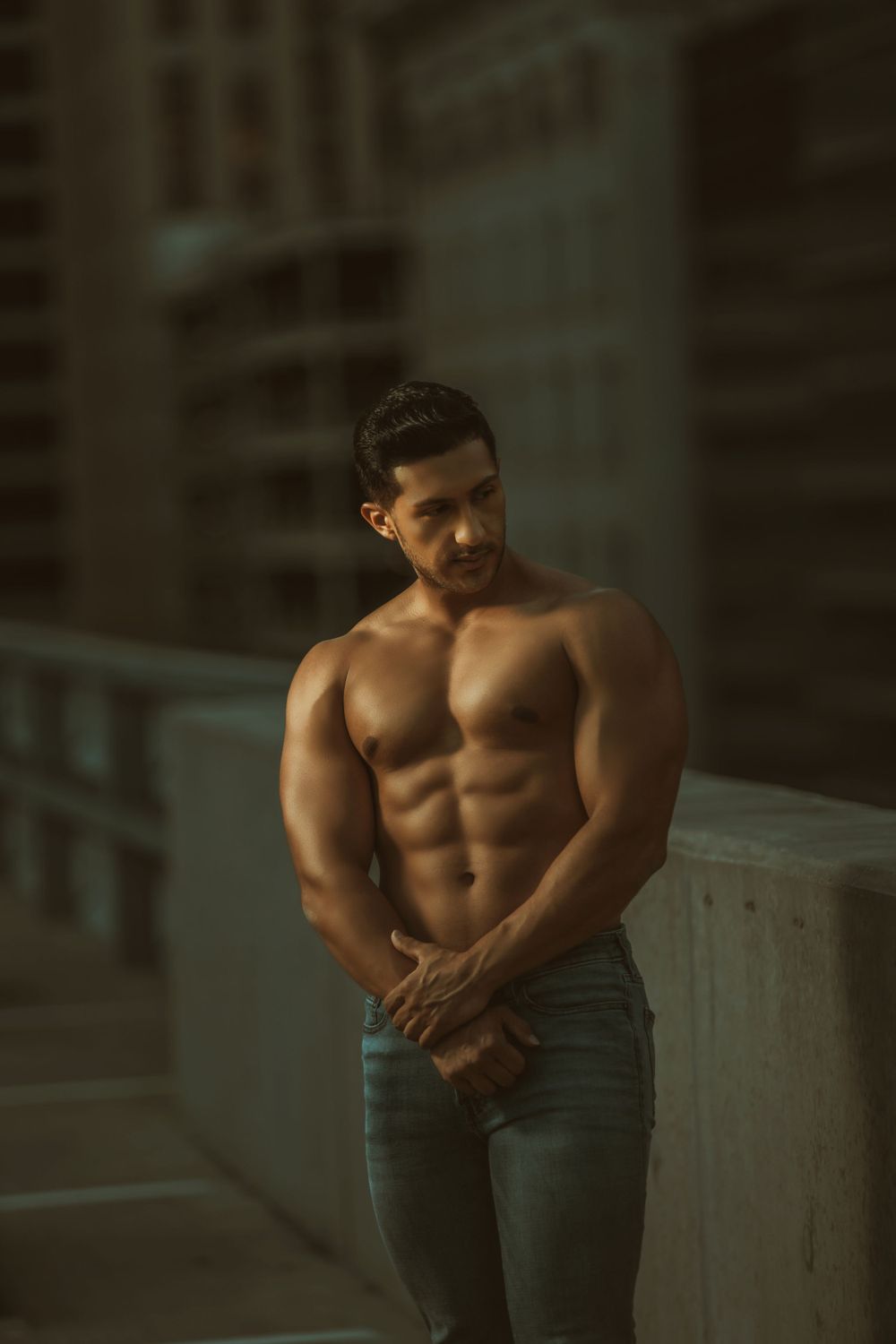 Athletic shirtless person in blue jeans poses against urban background in moody lighting.