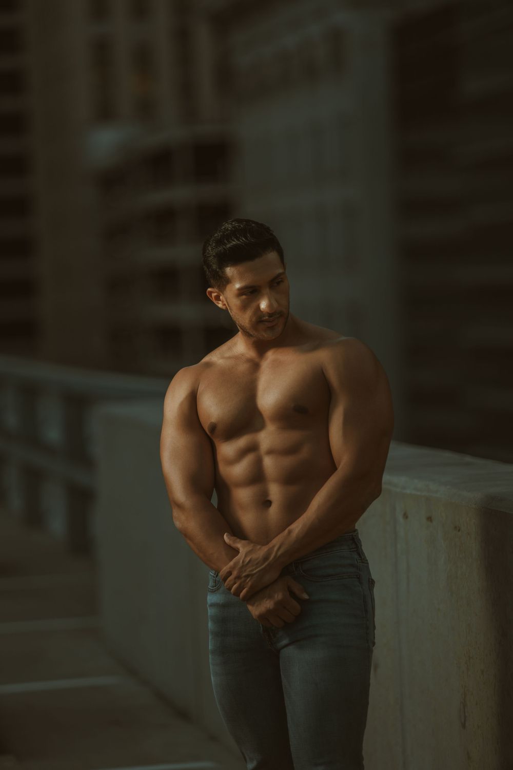 Muscular shirtless fitness model in jeans poses against urban backdrop in moody lighting.