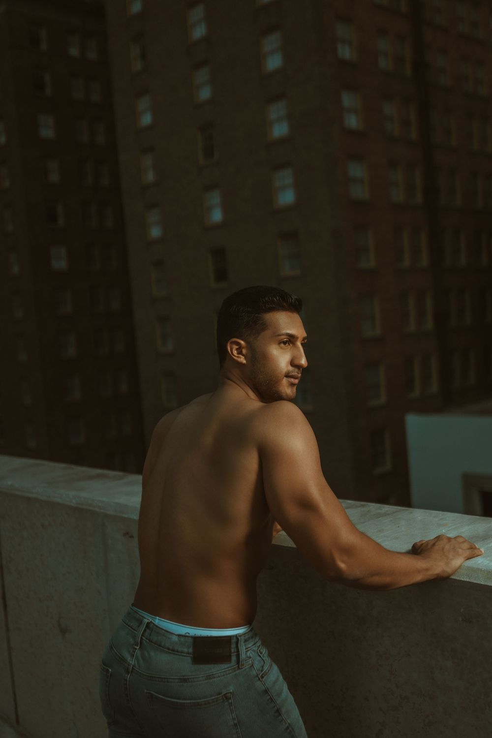 A muscular shirtless man in jeans leans against a city rooftop edge during an atmospheric golden hour shoot.
