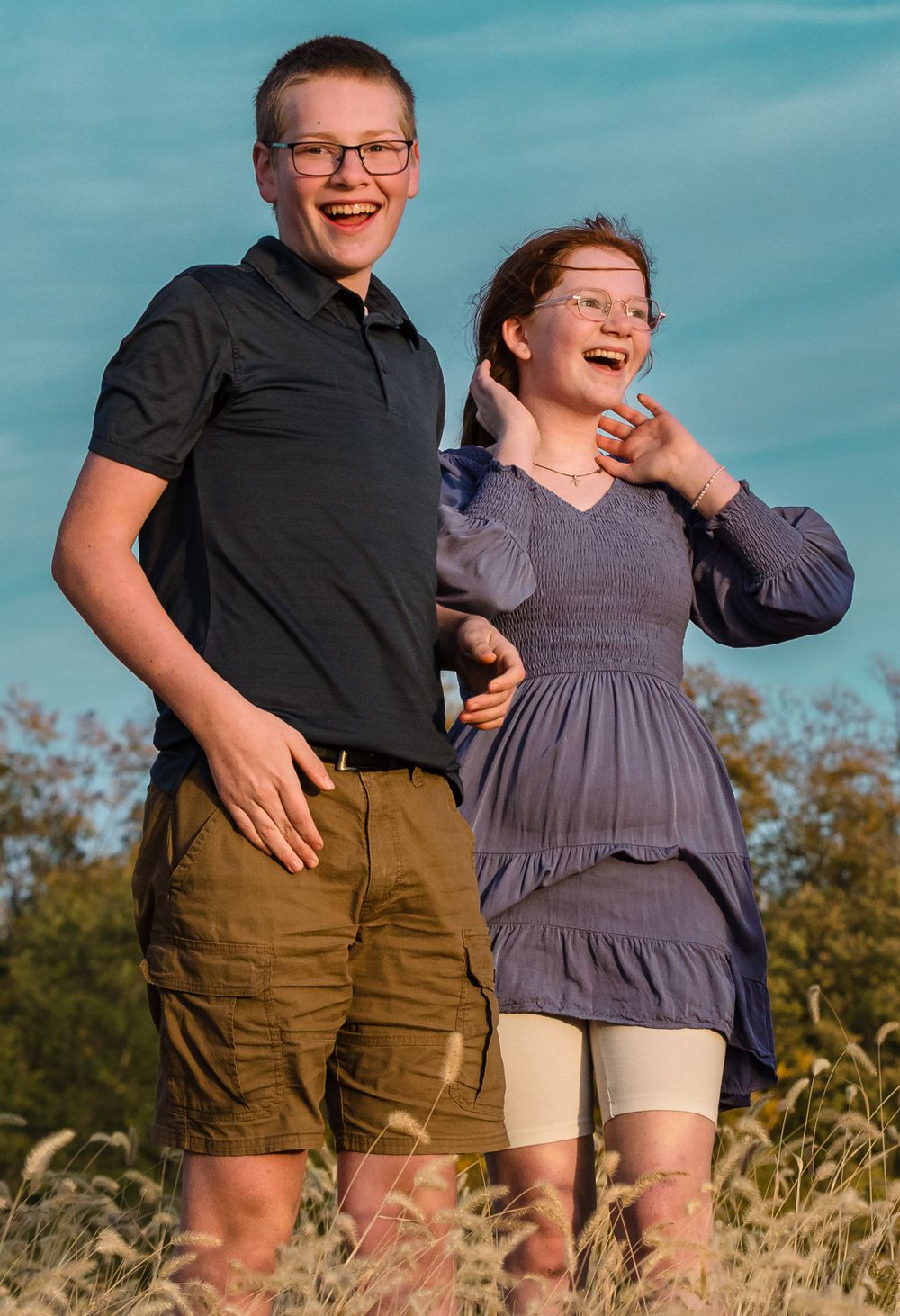 A joyful outdoor portrait of two people laughing together against a blue sky, wearing casual attire in a field.