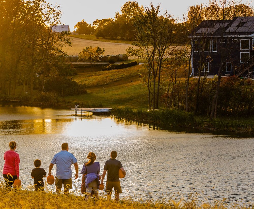 A group of people stand by a tranquil pond at sunset with golden light illuminating rolling hills and an old barn.