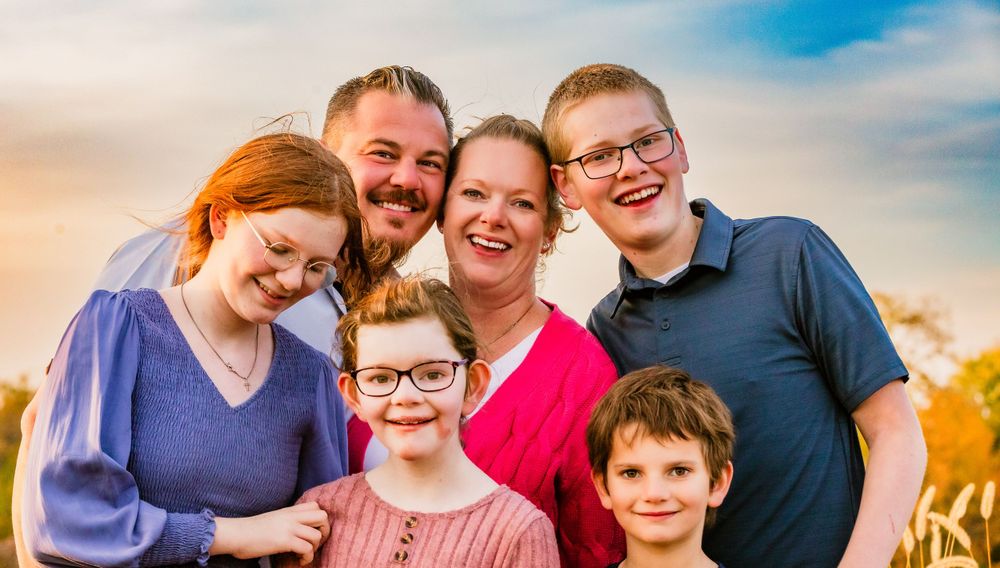 A group photo of a family of six smiling together outdoors against a sunset sky.