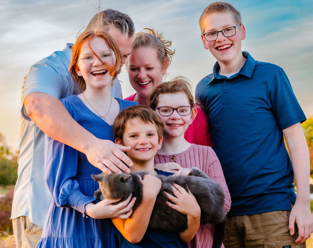 A cheerful family poses together outdoors holding a small gray cat against a sunset sky backdrop.