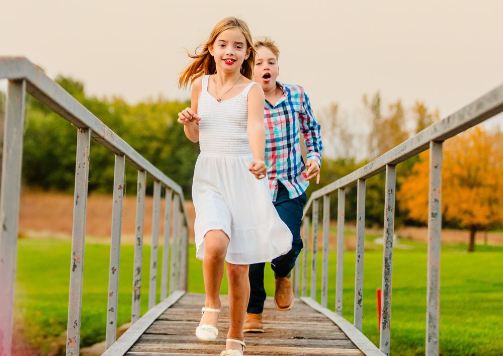 Two people running down a wooden bridge walkway surrounded by autumn trees and grassy fields on a sunny day.