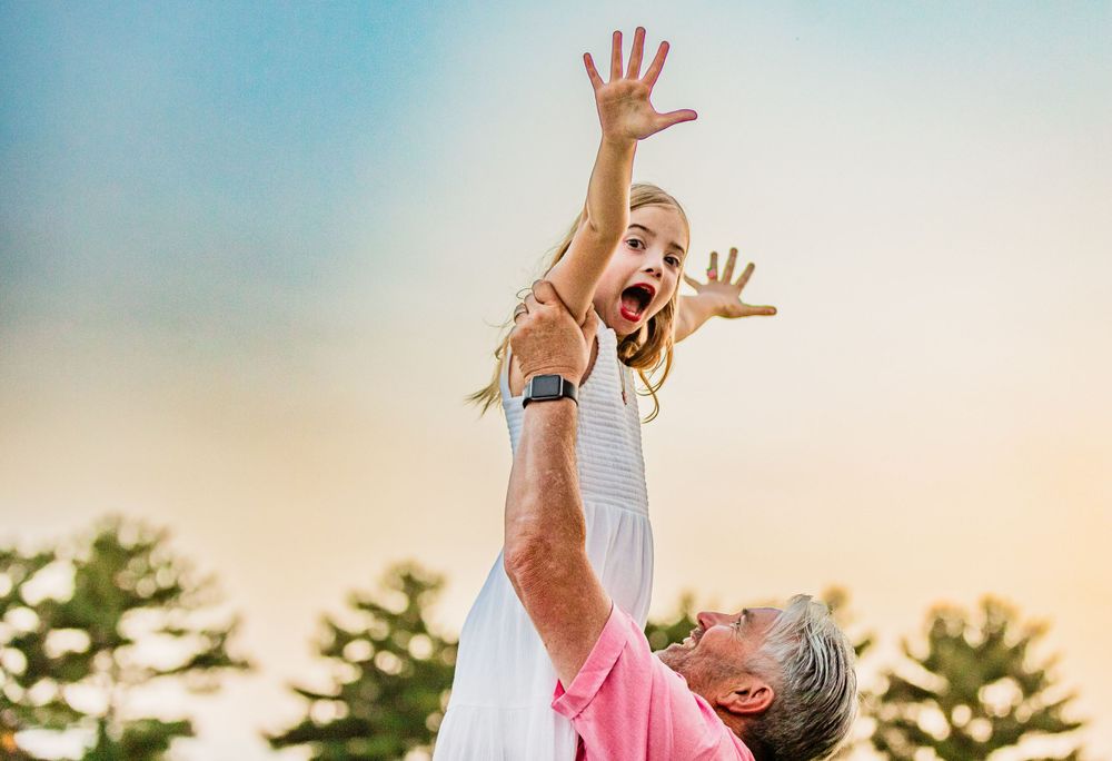 A joyful moment captured at sunset as someone lifts a child high into the air with outstretched arms.
