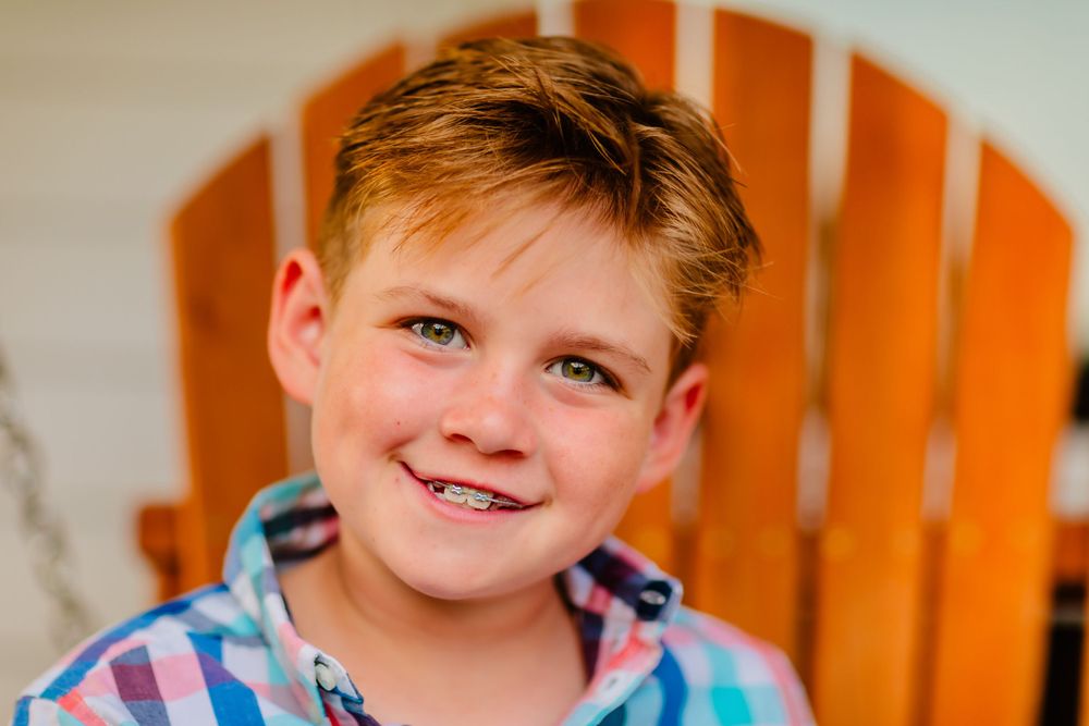 Child with spiky blonde hair wearing a plaid shirt poses in front of an orange wooden fence.