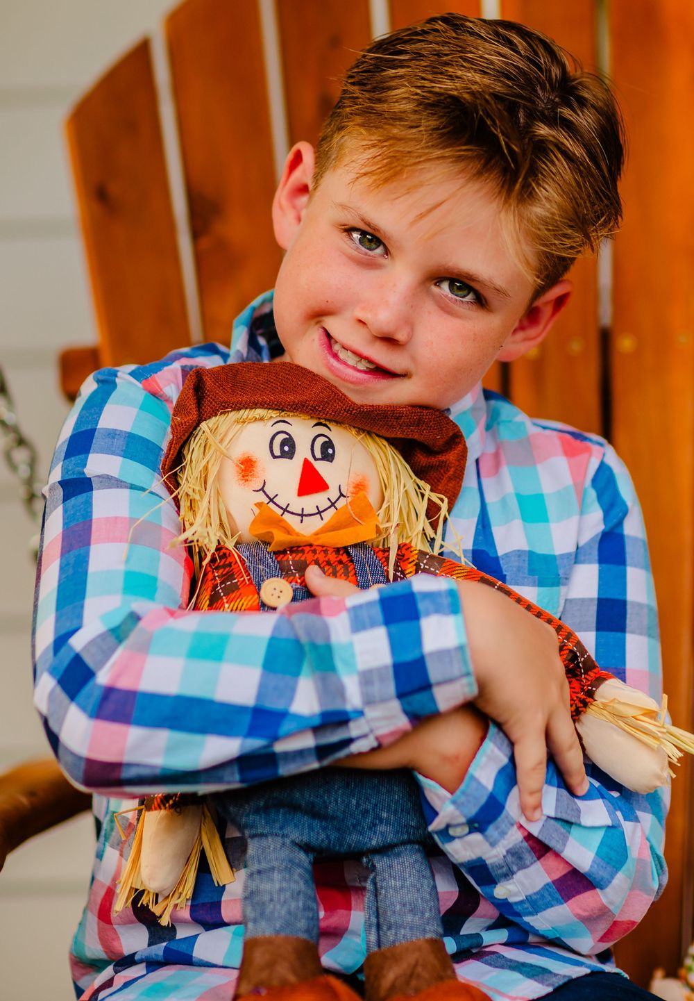Boy in blue plaid shirt hugging adorable scarecrow decoration with smiley face and straw hair.