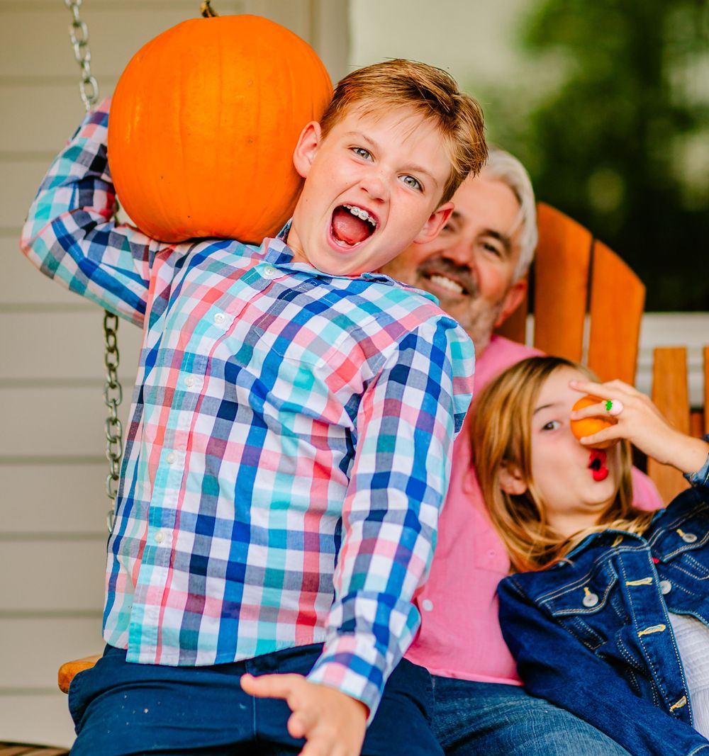 Family enjoying autumn fun with pumpkins on a porch swing, showing playful expressions and plaid shirt.