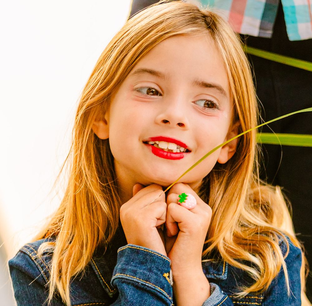 Young person with bright red lipstick and blonde hair wearing denim jacket smiles while posing outdoors.