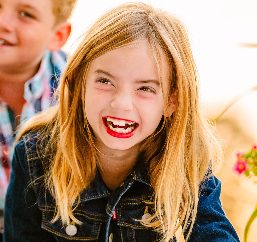 Joyful child with blonde hair laughs expressively while wearing a denim jacket in bright sunlight.