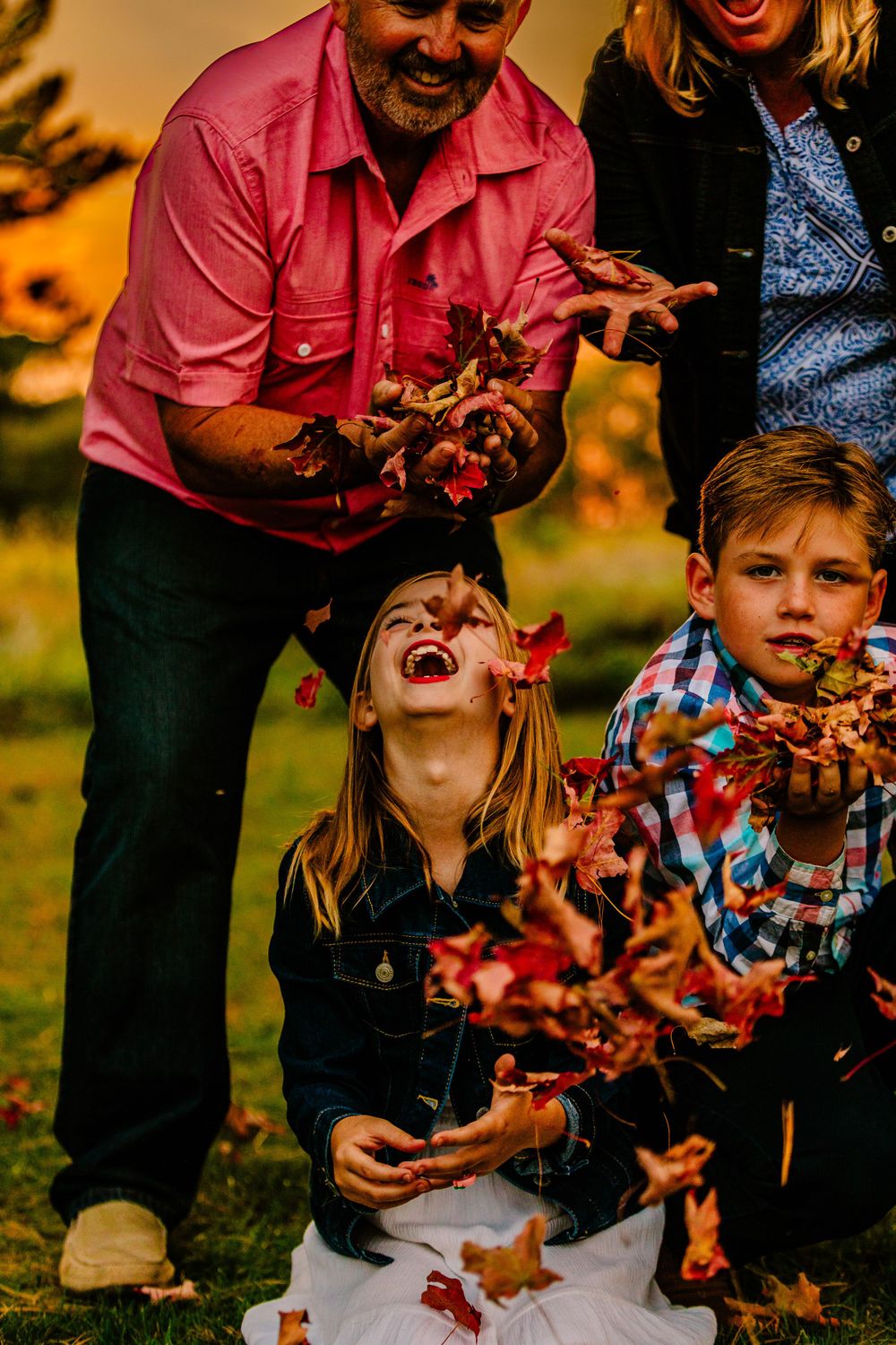 Family plays together outdoors throwing autumn leaves in the air during golden hour sunset light.