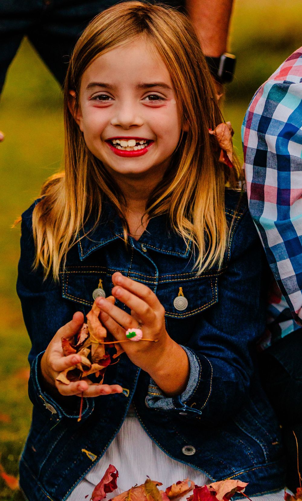 Someone wearing a denim jacket and white dress smiles while playing with flower petals outdoors.