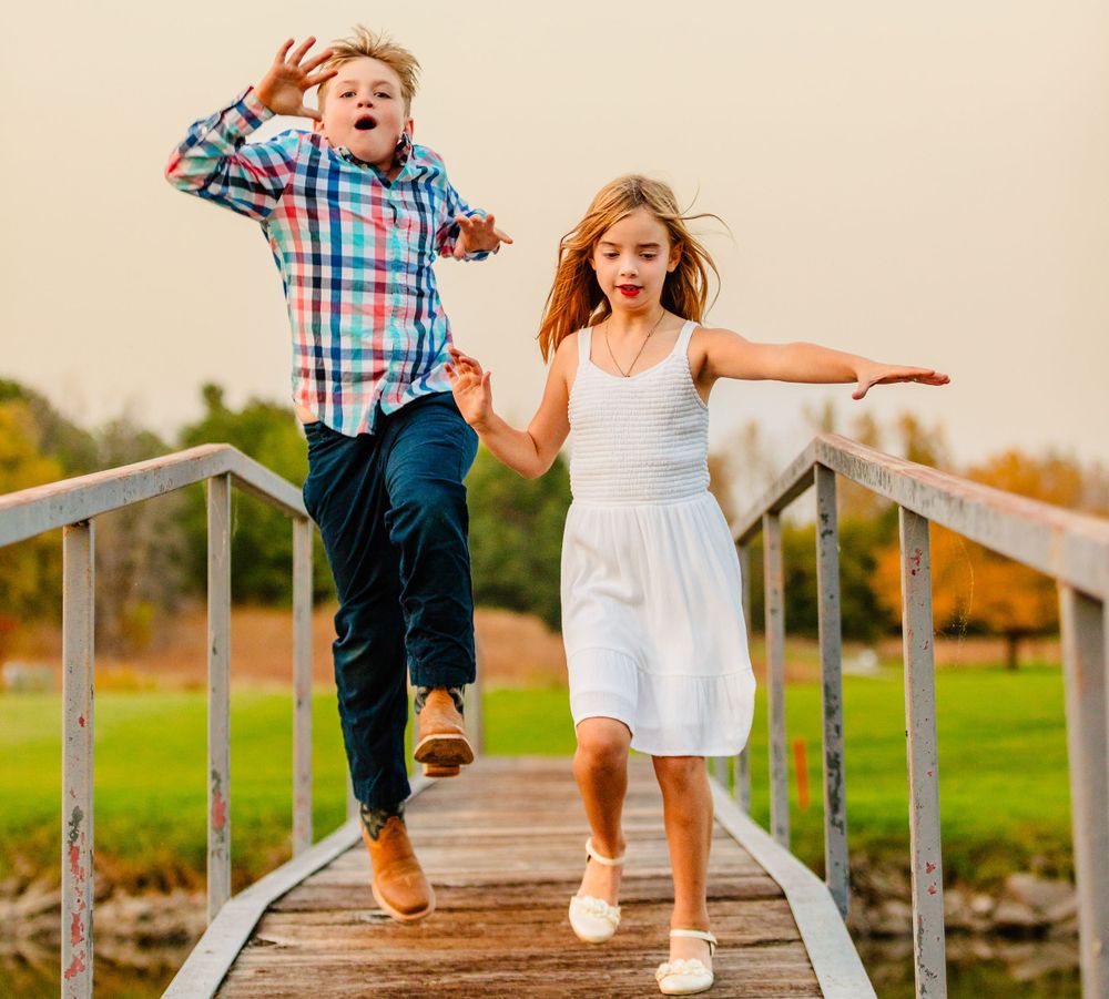 Two children jump playfully on a wooden bridge in a park during golden sunset light.