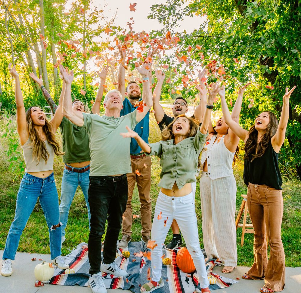 A group of friends joyfully throwing autumn leaves in the air at a park during sunset while standing together.