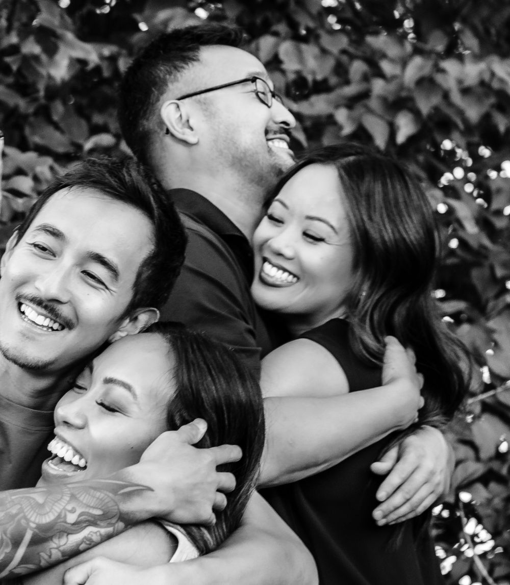 Black and white candid photo showing a joyful group embrace with genuine laughter and happiness against a leafy background.