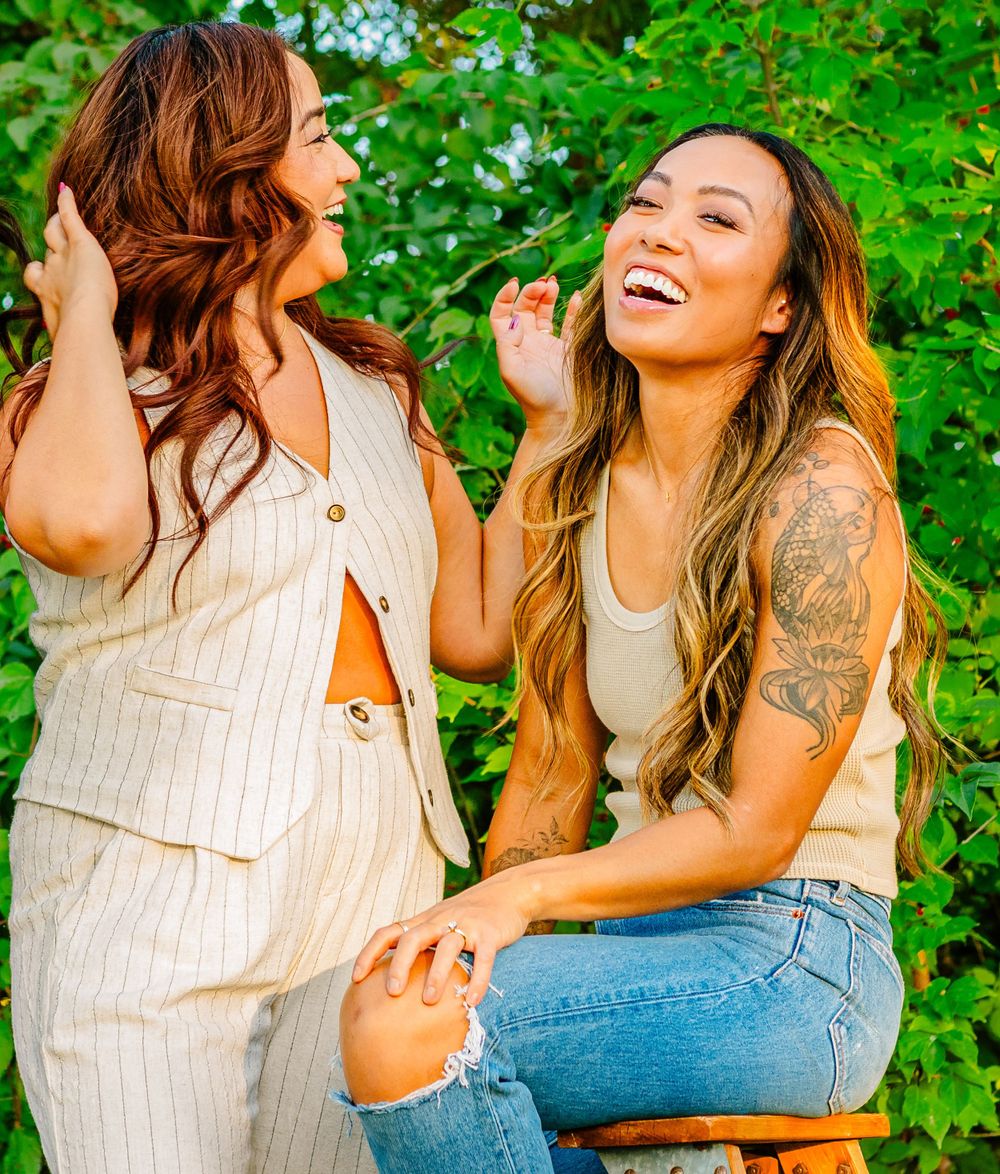 Two friends share a joyful moment laughing together outdoors against green foliage background.