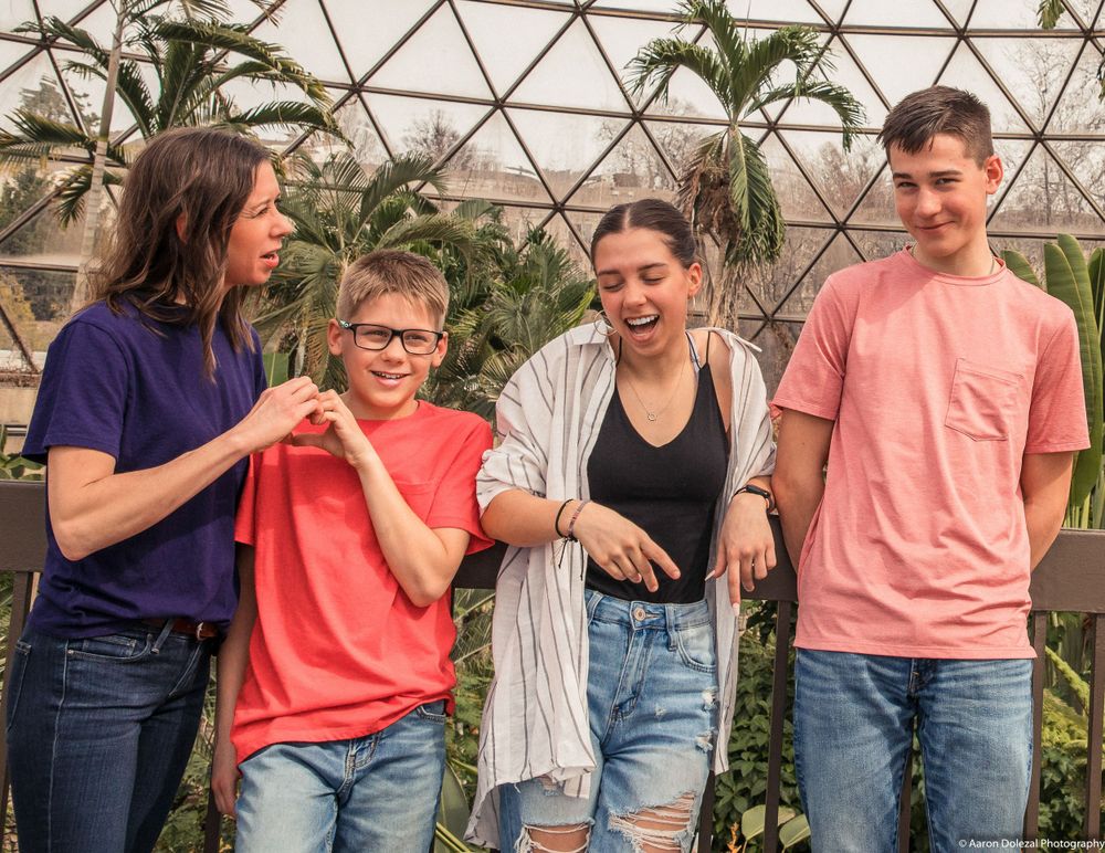 Four people in casual clothing stand together laughing and smiling in front of plants inside a greenhouse dome structure.