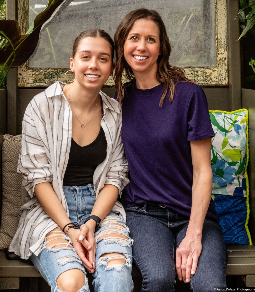 Two people in casual attire sitting together on a bench with floral cushions in an outdoor setting.
