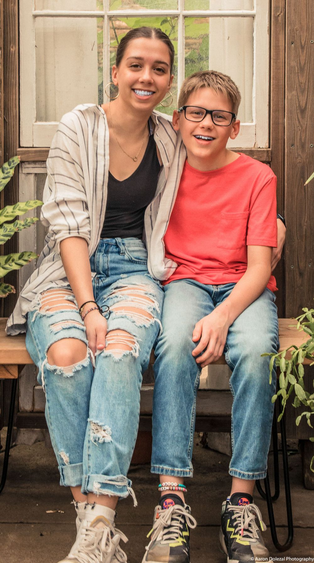 Two people sitting together on a porch wearing casual jeans and sneakers with a wooden fence in the background.