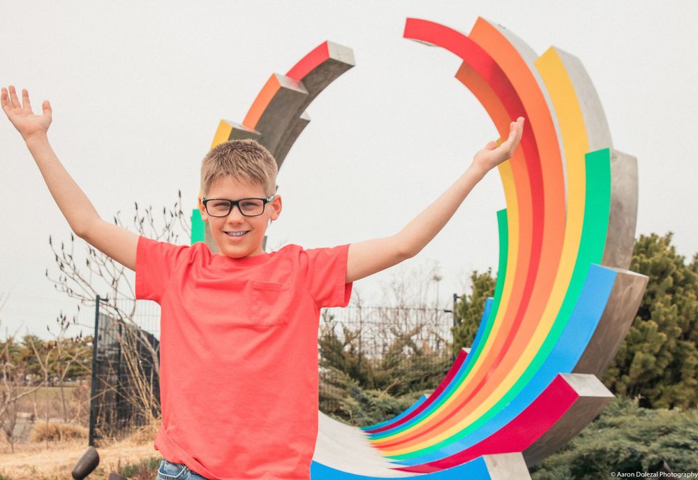 A joyful person in a coral t-shirt jumps with raised arms in front of a colorful rainbow-shaped outdoor art installation.