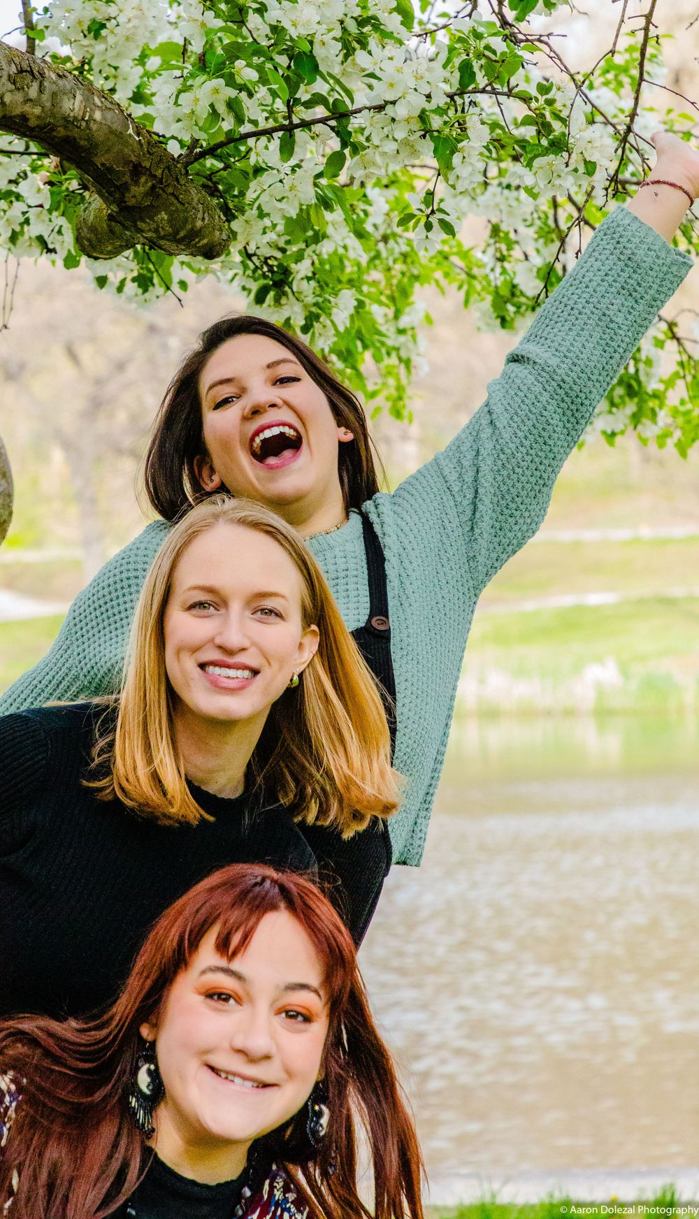Three friends enjoying a joyful moment together under a tree with green spring leaves by a lake.