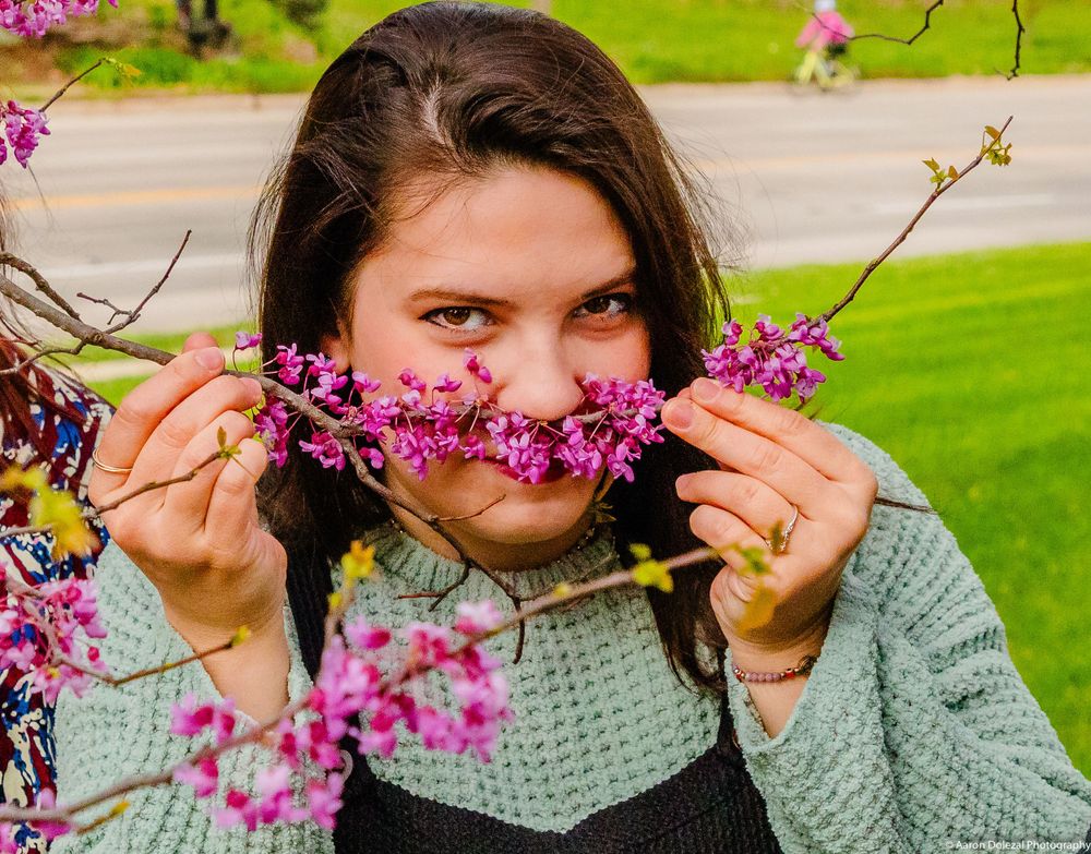 Someone in a mint green sweater poses playfully with pink redbud tree blossoms in a spring garden setting.