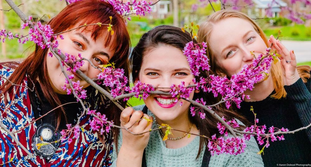 Three people joyfully pose with vibrant purple redbud tree blossoms in front of them during springtime.