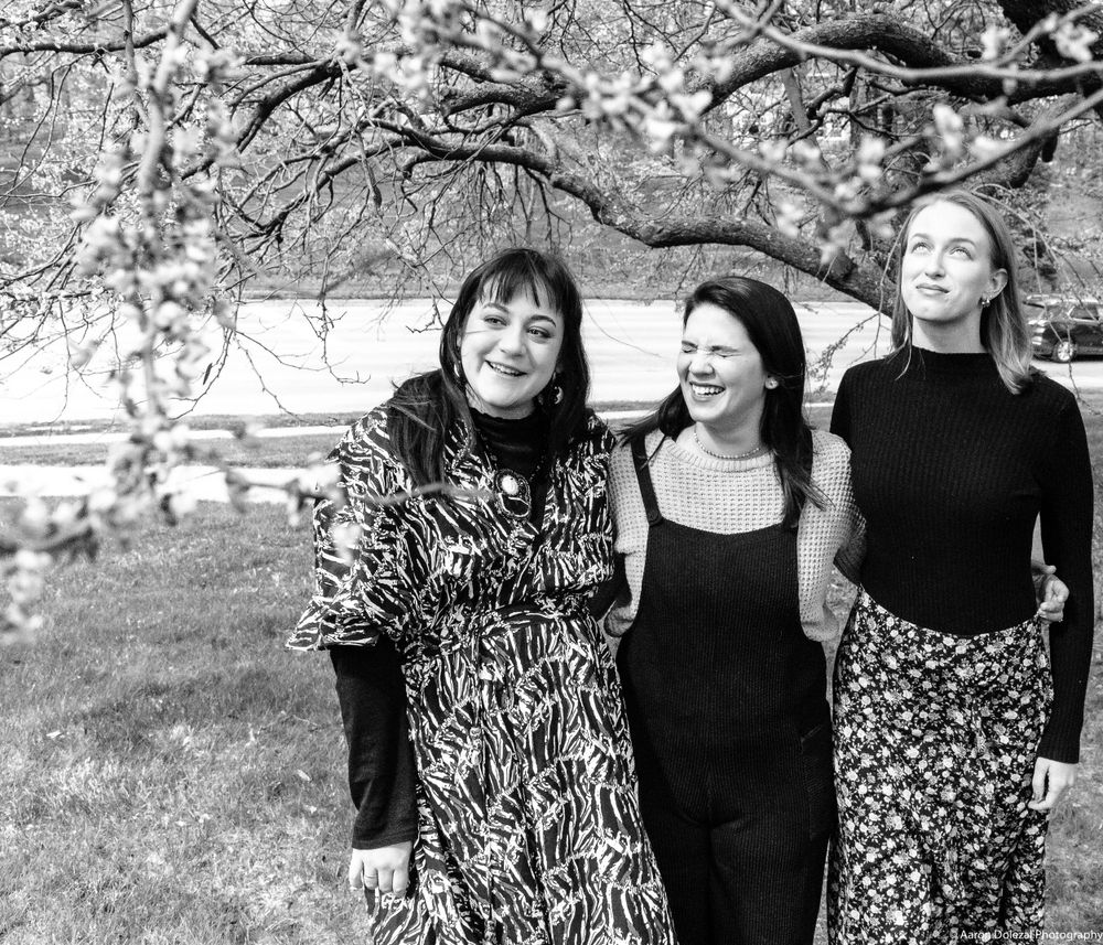 Three women in black and patterned dresses pose together under blooming cherry blossoms in a black and white photo.