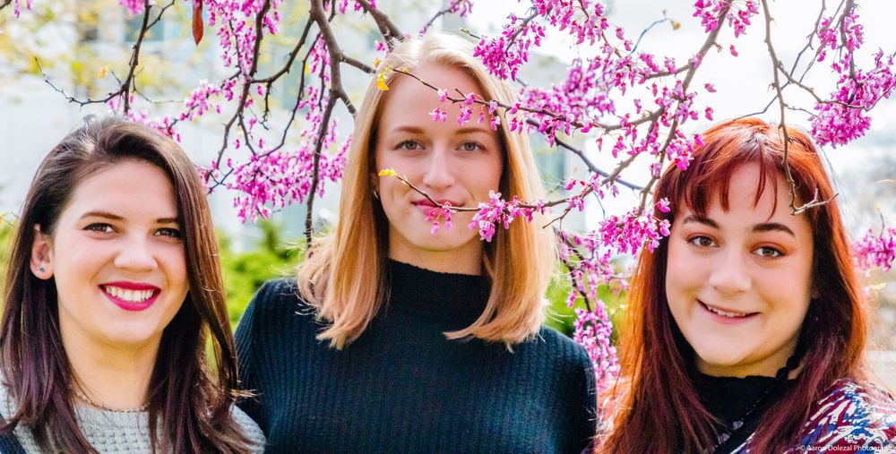 Three women in black tops smile while posing together under pink spring cherry blossoms.