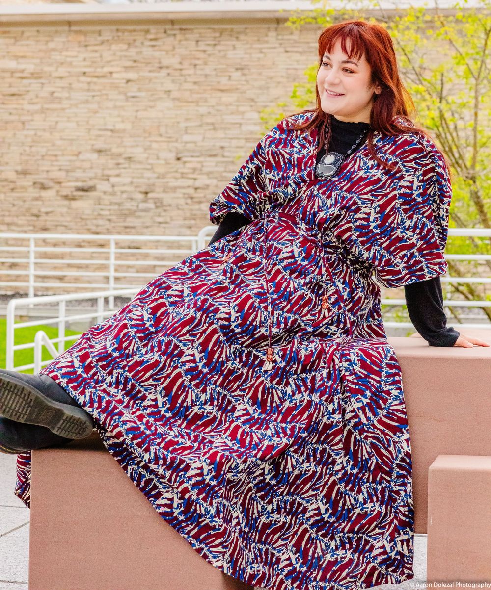 A model in a pink and blue patterned dress with long sleeves sits on a concrete ledge outdoors.