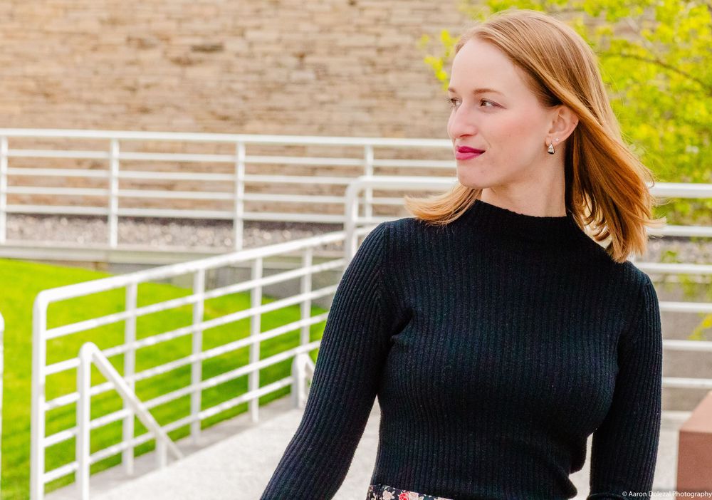 Person wearing a black ribbed sweater and red lipstick poses outdoors near white fencing on a sunny day.