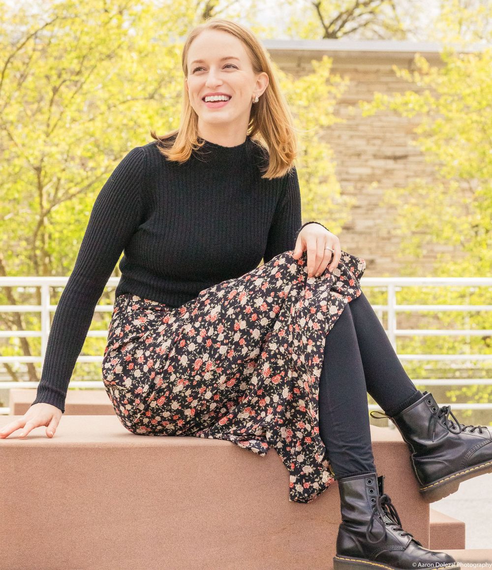 Someone wearing a black turtleneck sweater and floral skirt sits casually on a ledge with autumn foliage in the background.