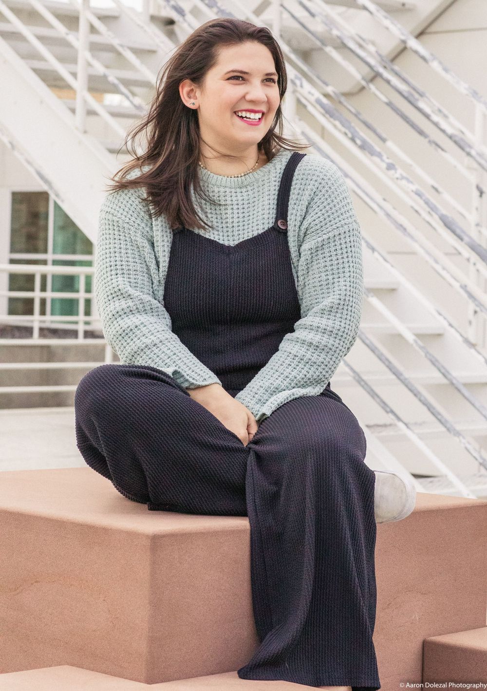 A person wearing a cozy gray sweater and black overalls sits casually on outdoor steps with a warm smile.