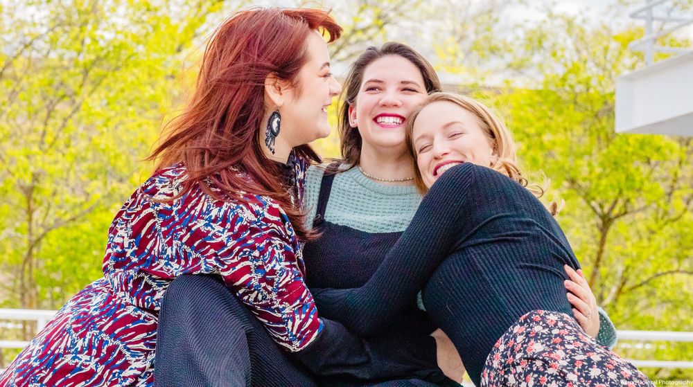 Three people share laughter and warm embrace while sitting outdoors with yellow spring blossoms in the background.