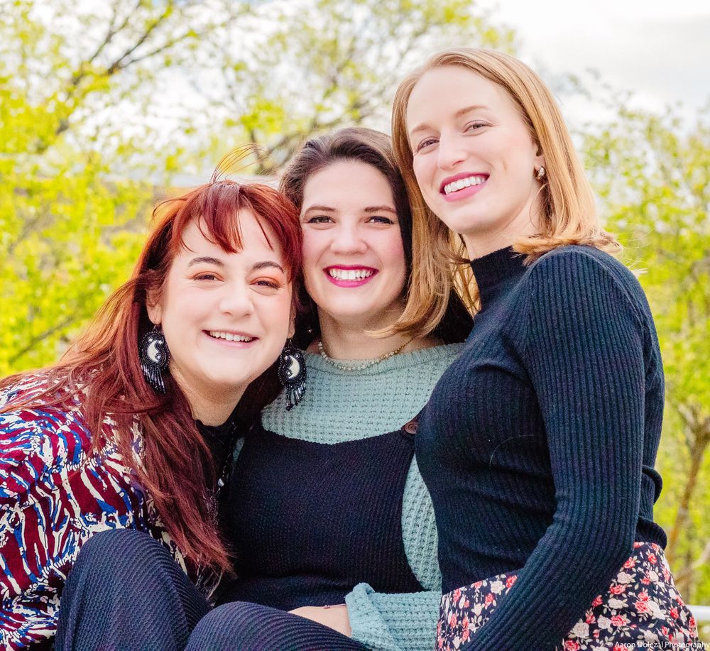 Three friends sitting together outdoors on a sunny day with trees and yellow flowers in the background.