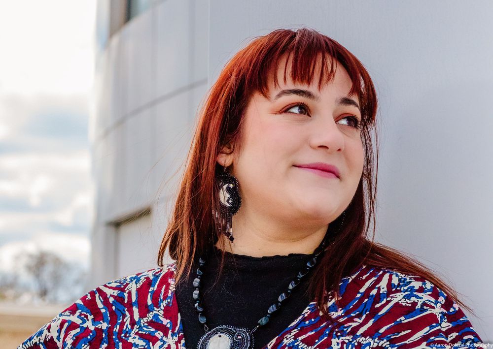 Person wearing a colorful patterned cardigan and black necklace smiling while looking up at the sky outdoors.