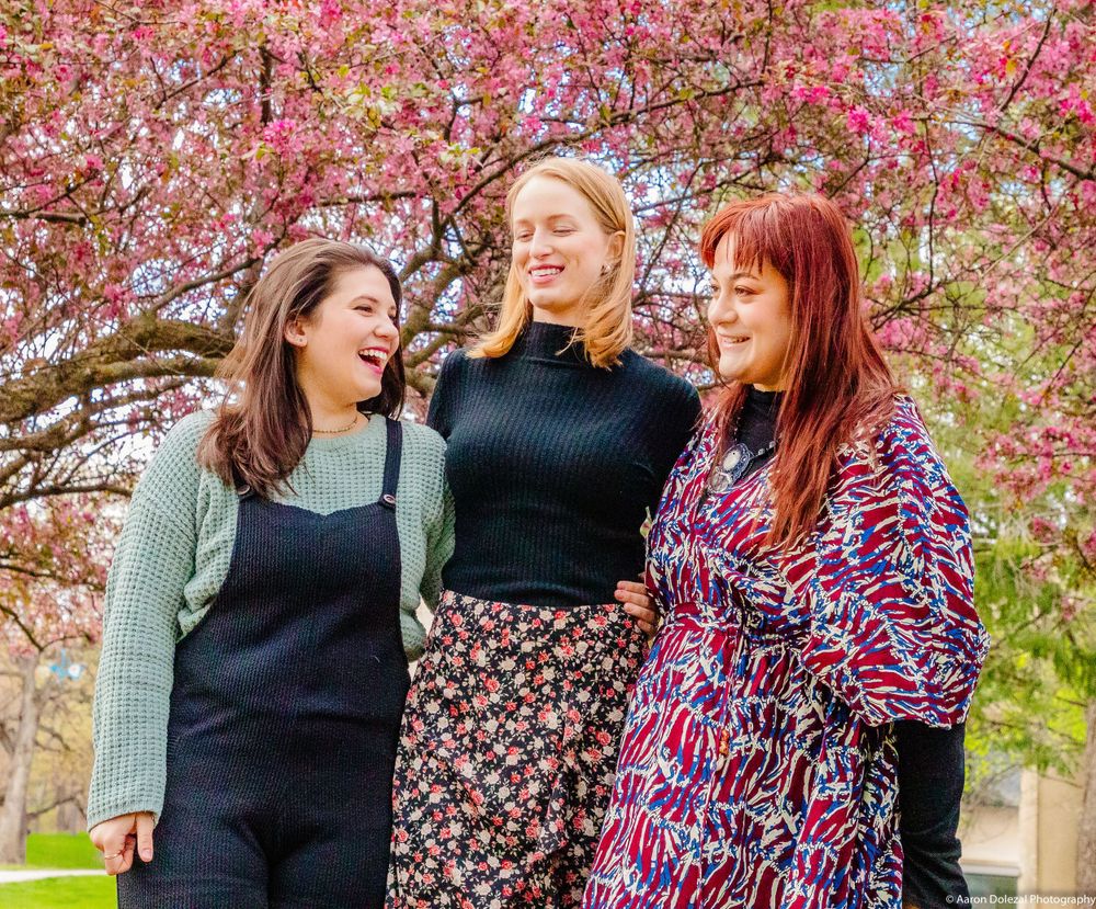 Three friends posing together in front of blooming cherry blossom trees during springtime.