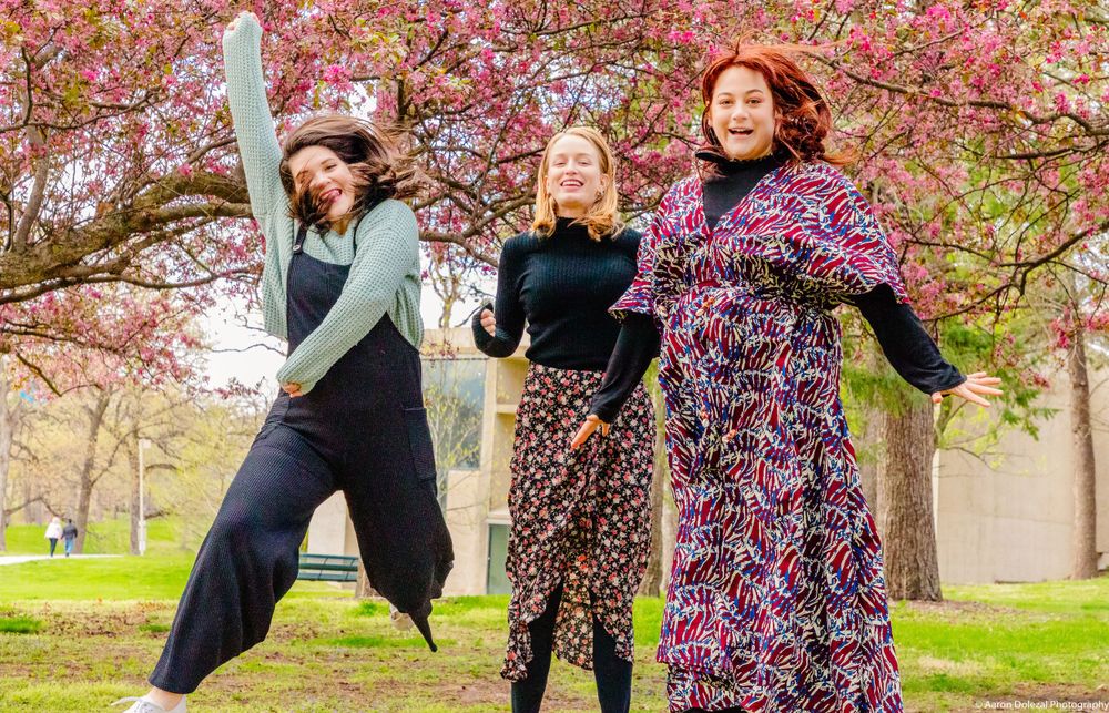 Three friends dancing joyfully under pink cherry blossom trees in a park.