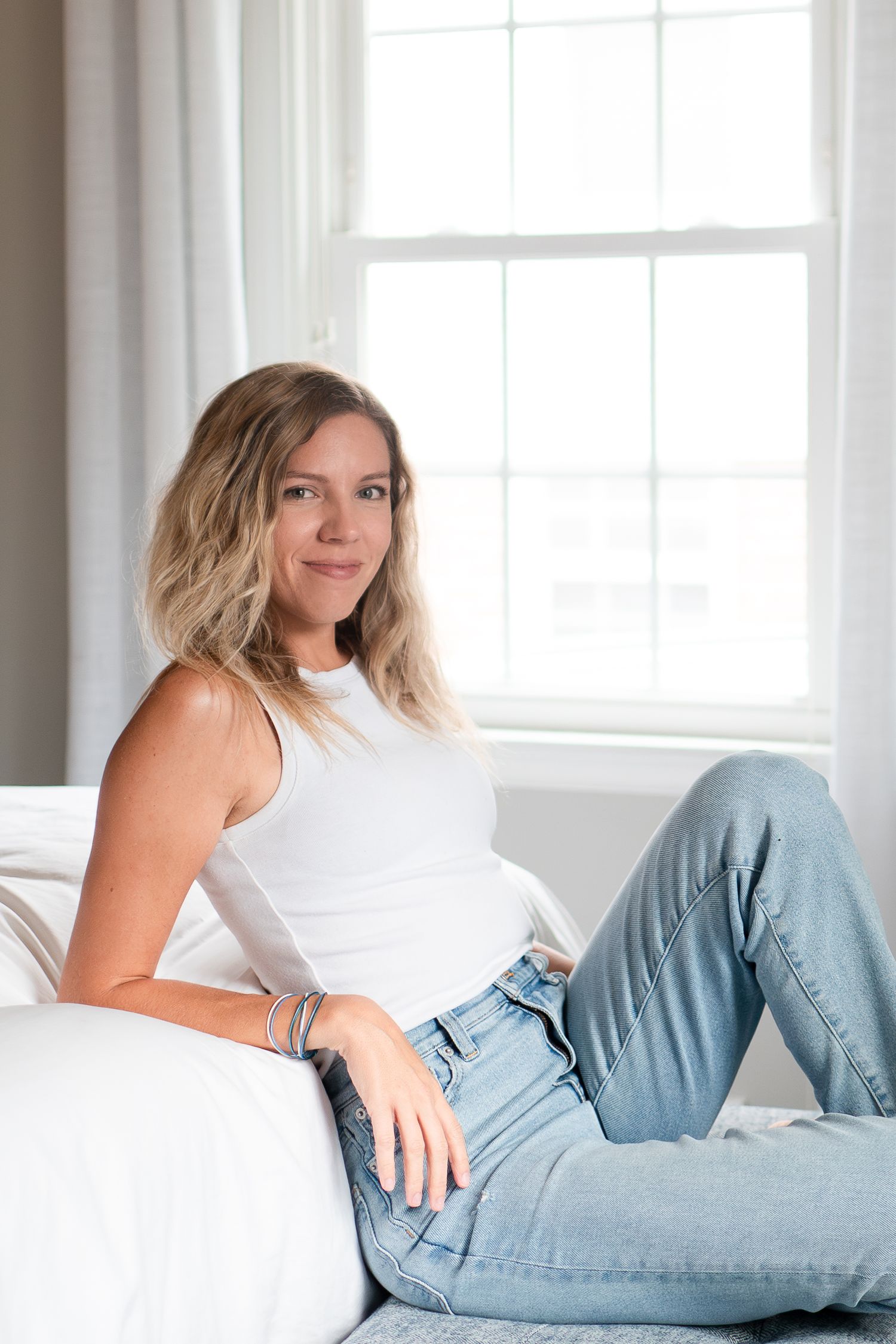 A casual lifestyle portrait of someone relaxing on a white couch wearing a white tank top and light wash jeans.