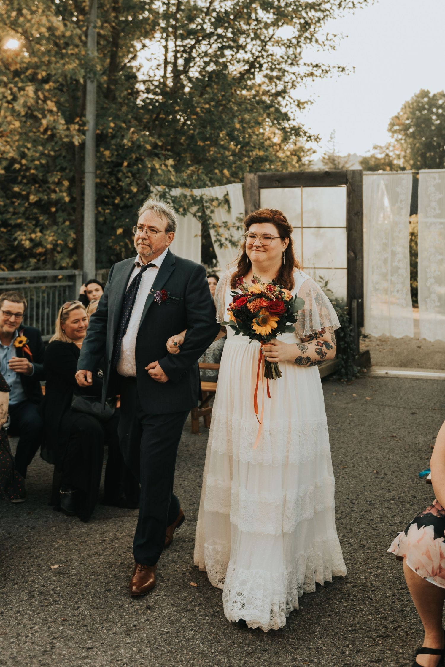 A bride in a vintage lace wedding dress walks with her escort while carrying a vibrant autumn bouquet at an outdoor ceremony.