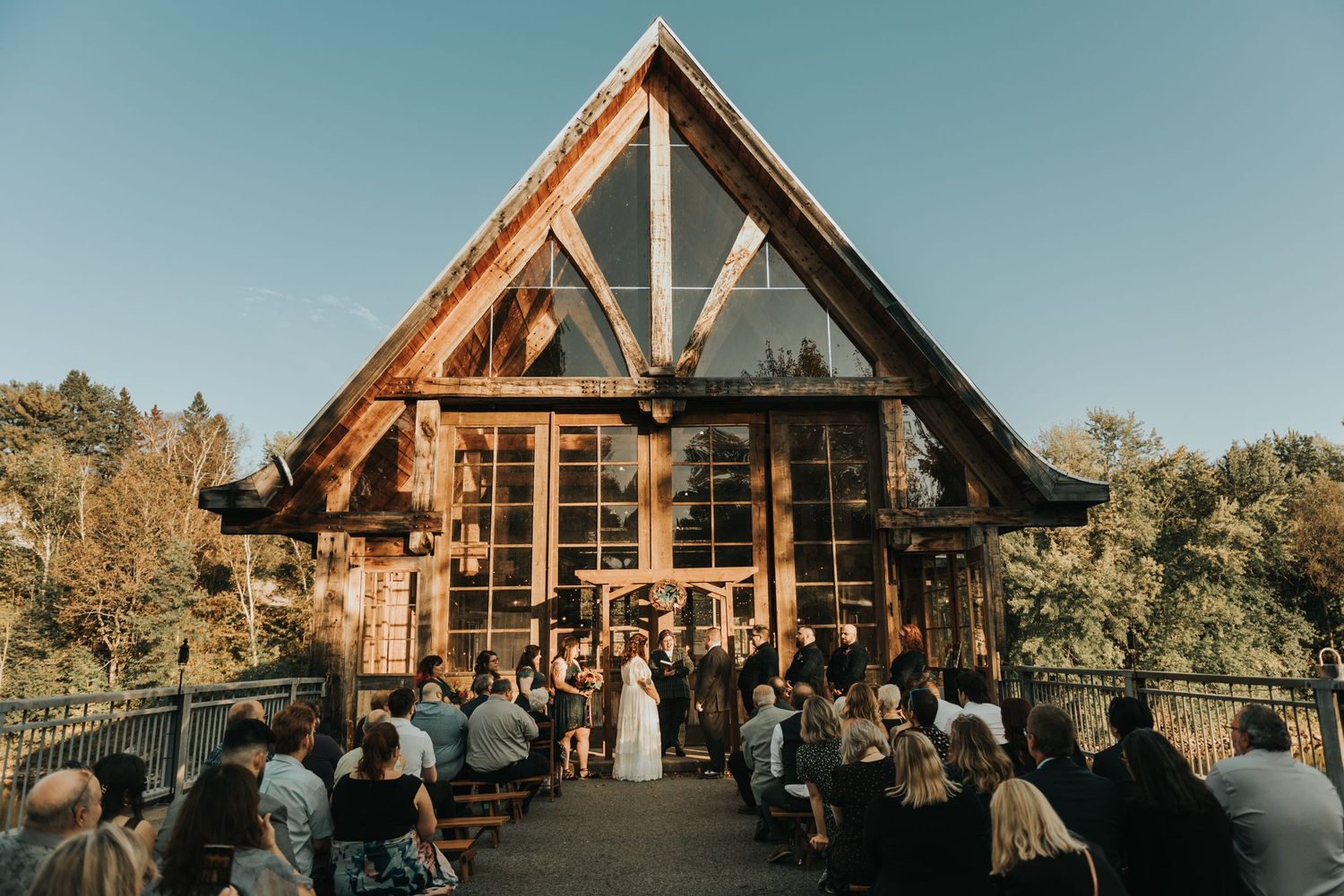 Golden hour wedding ceremony at a modern wooden lodge with A-frame architecture and panoramic glass windows.