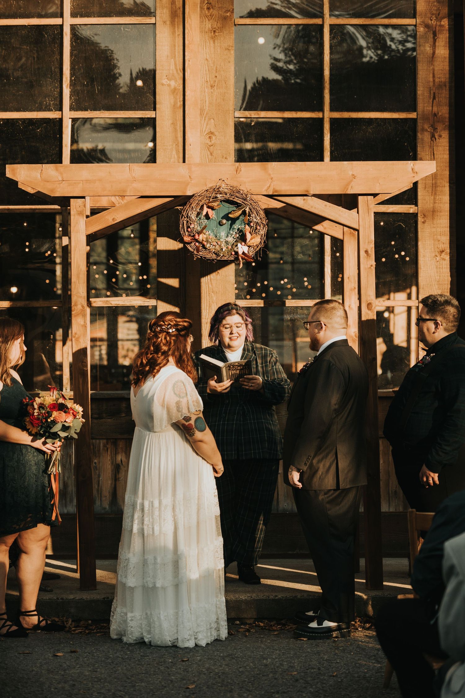 Wedding ceremony takes place in front of large windows at sunset with rustic wooden pergola and decorative wreath.