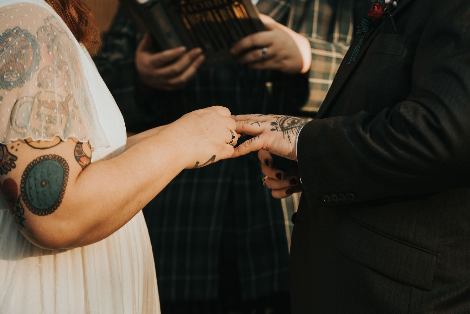 Close up of hands during ring exchange at wedding ceremony showing sleeve of white dress and tattooed arm.