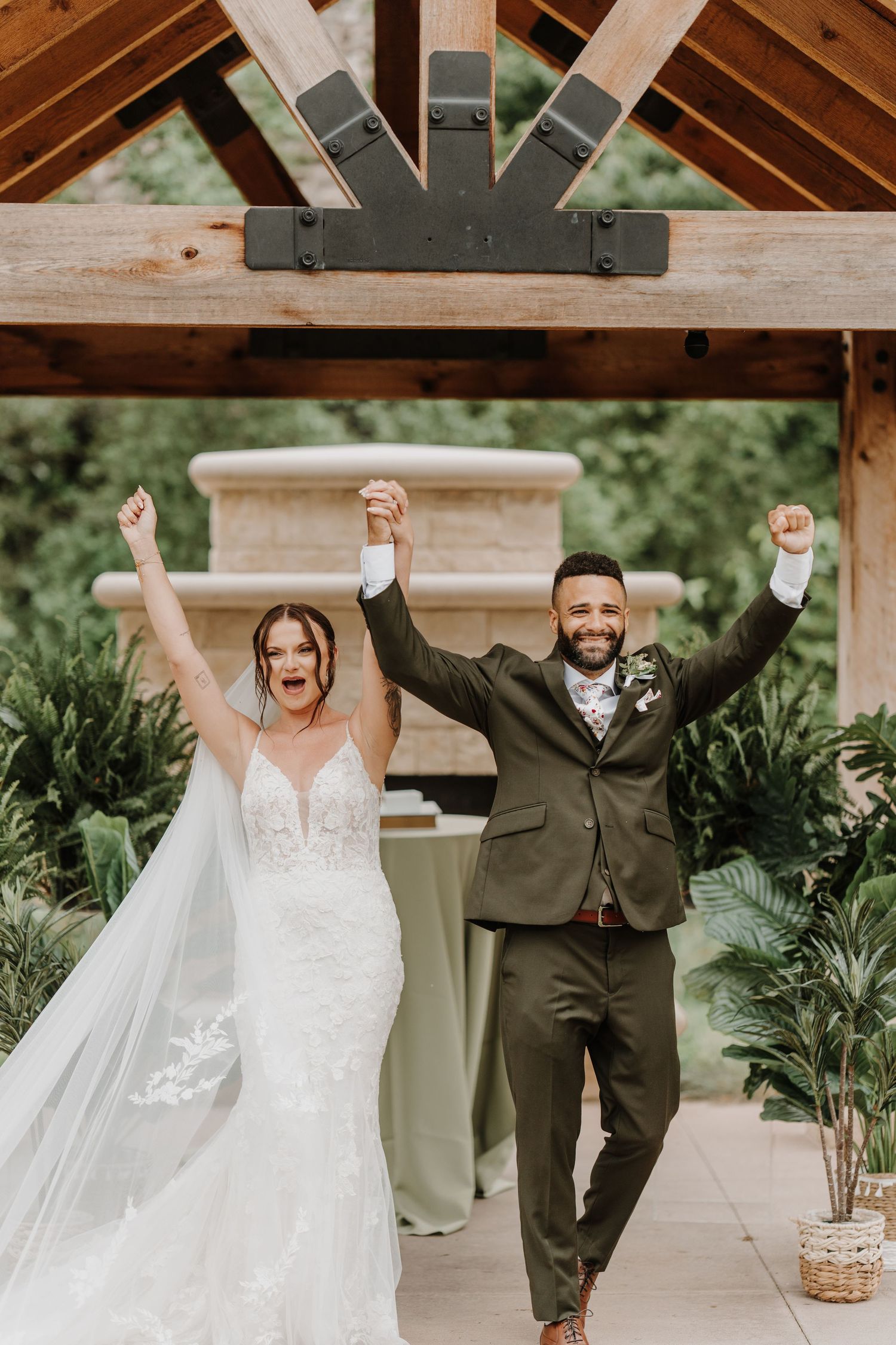 Joyful couple celebrates during their wedding under a wooden overhang in a lush green outdoor setting.