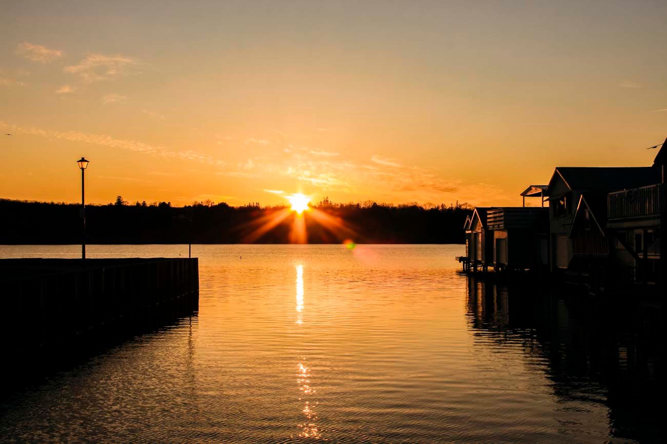 Golden sunset over calm harbor waters with silhouetted dock structures and a sunburst reflecting on the water's surface.